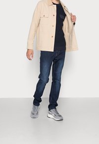 Beige bomullsjacka med två bröstfickor, marinblå tröja under, mörkblå jeans och grå sportskor med vita sulor.