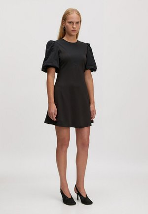 Schwarzes kurzes Kleid mit Puffärmeln, enganliegendem Oberteil und schwingendem Rock. Glattes Material mit strukturiertem Ärmelabschluss. Schwarze High Heels.