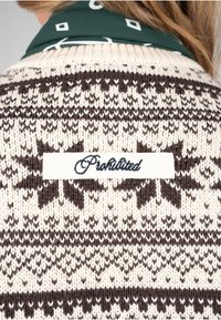 Gros plan d’un pull en tricot beige et marron avec un patch blanc brodé du mot "Prohibited" dans le dos.