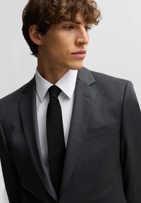 Grijs op maat gemaakt pak met een enkele zak, gecombineerd met een witte dress-shirt en zwarte stropdas. De stof heeft een gladde textuur en een slank ontwerp.