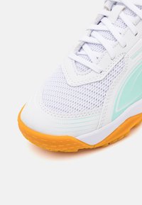 Hvit atletisk sko med overdel i mesh, myntegrønne detaljer og en strukturert oransje såle for grep. Snøring integrert i designet.