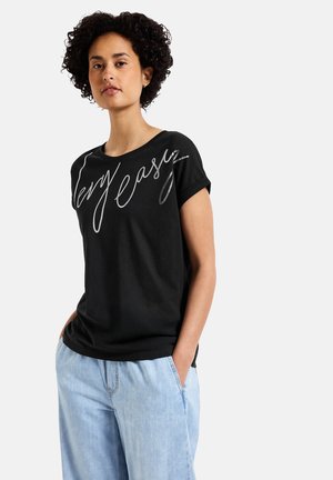 Camiseta negra de manga corta con escote redondo y texto en cursiva plateada "Muy fácil" en la parte frontal. Se lleva con pantalones holgados de color azul claro.