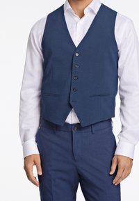 Námořnický modrý vest v hladkém materiálu, se stojáčkem do V, šesti knoflíky a dvěma předními kapsami, spárován s bílou košili.