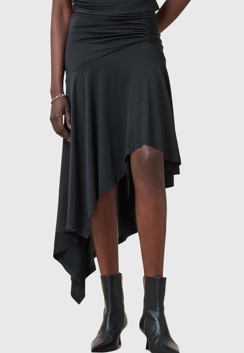 Femme portant une jupe asymétrique noire avec taille plissée et bottines noires à fermetures éclair latérales, debout devant un fond uni.