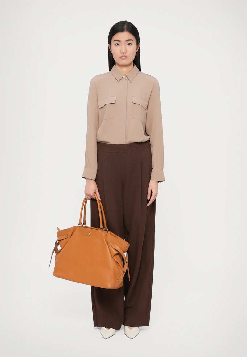 Brūns ādas rokassomas ar apaļu formu, divām rokturiem un dekoratīvām sānu detaļām. Modelis valkā bēšu krāsas garām piedurknēm blūzi un brūnas plašas bikses.