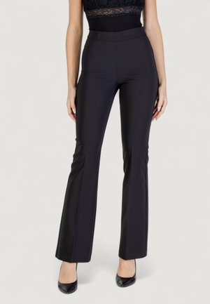 Donna con pantaloni a palazzo neri a vita alta e tacchi neri, in piedi su uno sfondo neutro.