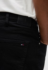 Κοντινό πλάνο ενός ατόμου που φορά μαύρο τζιν και λευκό πουκάμισο, εμφανίζοντας την πίσω τσέπη με μια μικρή λεπτομέρε ια σημαίας σε κόκκινο, άσπρο και μπλε.