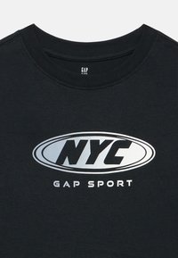 Tricou negru din bumbac cu decolteu rotund, având un logo mare argintiu "NYC" și text mai mic "GAP SPORT" dedesubt.