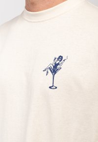 Bavlněné tričko v krémové barvě s modrou grafikou ženy sedící v koktejlové sklenici, která drží drink a cigaretu.