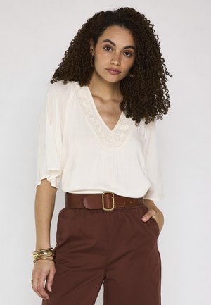 Donna con capelli ricci indossa una camicetta bianca con scollo a V, pantaloni marroni a vita alta, una cintura larga marrone e braccialetti dorati, posando su uno sfondo semplice.