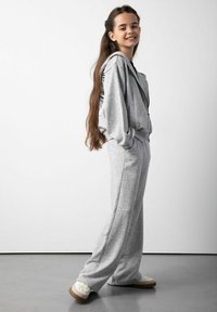 Szara bluza z kapturem i pasujące dresy wykonane z miękkiego materiału, o luźnym kroju i zamkiem z przodu. Model nosi białe trampki.