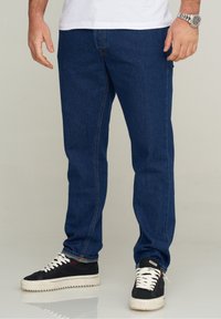 Vaqueros de mezclilla azul oscuro con corte recto, costuras visibles y diseño de cinco bolsillos. Llevados con zapatillas negras con cordones blancos.