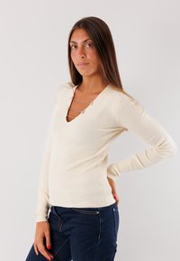 Maglione a coste di colore crema con scollo a V e dettagli in metallo decorativi all'escollatura. Maniche lunghe e silhouette aderente.