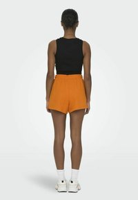 Zwarte mouwloze top en oranje high-waisted shorts. De shorts hebben een relaxed fit en een elastische tailleband, gecombineerd met witte sokken en sneakers.