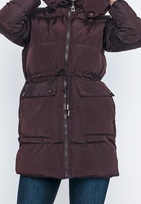 Henkilö pukeutuneena tumman marjaiseen tikattuun toppatakkiin, jossa on etuvetoketju, säädettävä vyötärönauha, nappikiinnitteiset taskuklapit ja kohotettu kaulus.