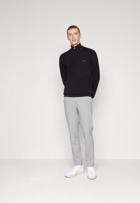 Zwarte half-zip pullover van zachte stof, gecombineerd met lichtgrijze uitstraling en witte sneakers, wat zorgt voor een moderne, minimalistische look.
