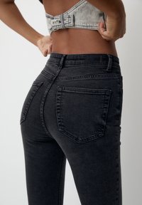 Jean en denim noir à coupe slim avec poches arrière, porté par une personne ajustant un haut en denim gris clair, coupé court.