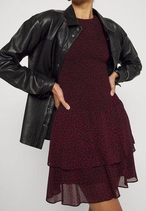 Kobieta w czarnej skórzanej kurtce na czerwono-czarnej sukience w panterkę z marszczonym stanikiem i warstwową spódnicą, ręce na biodrach.