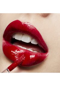 Gloss à lèvres rouge appliqué sur les lèvres, mettant en valeur une finition brillante et une texture lisse. Des dents blanches sont visibles, renforçant le contraste de couleur éclatant.