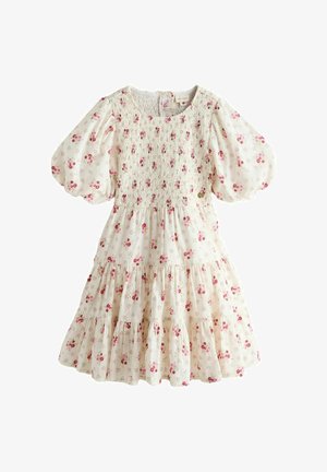 Fiatal lány mosolyogva, előre sétálva, fehér ruhát visel, apró piros virágmintákkal és bézs szandállal, egyszínű háttér előtt.