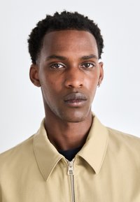 Chaqueta beige con cremallera y cuello puntiagudo, que presenta una textura suave y un diseño minimalista. Acentuada con detalles de costura sutil.