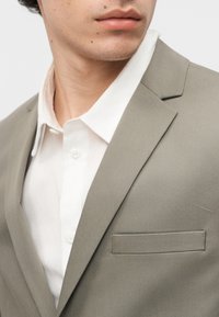 Κοντινό πλάνο ενός ατόμου που φοράει ένα ανοιχτό γκρι σακάκι πάνω από ένα λευκό πουκάμισο με κουμπιά, εστιάζοντας στην περιοχή του γιακά και του πέτο.