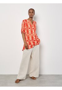 Rote Tunika mit geometrischen Mustern, V-Ausschnitt und kurzen Ärmeln, kombiniert mit beigen, weit geschnittenen Leinenhosen und braunen flachen Sandalen.