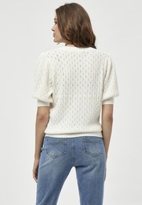 Wei�ßer gestrickter Pullover mit strukturiertem Muster, puffigen Ärmeln und geripptem Saum. Über blauen Jeans getragen, was einen lässigen Stil zeigt.