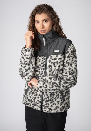 Woman with curly hair wearing a zipped, leopard-print fleece jacket with black shoulders and SBVL branding, posing against a plain background.