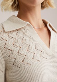 Svetlo bež pleten kardigan z V-izrezom in ovratnikom, ki se ponaša z zapletenimi vzorčastimi detajli in mehko teksturo. Vidna preprosta srebrna verižica.