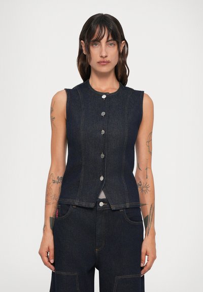 Kvinde med mørkt hår iført ærmeløs mørk denim skjorte med knapper og matchende jeans, stående mod en ensfarvet lys baggrund.