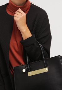 Svart läderhandväska med en rektangulär gyllene detalj, slät textur och dubbla handtag. Bärs med en svart cardigan över en rostfärgad topp.