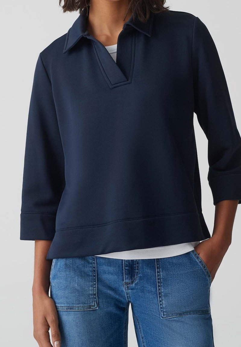 Femme portant un haut bleu marine à manches longues avec col en V par-dessus une chemise blanche, associé à un jean bleu, avec une main dans la poche.