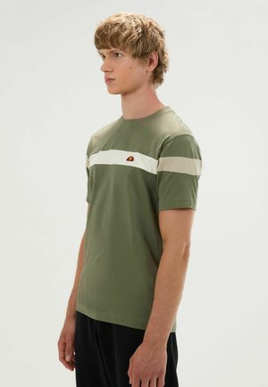 Junger Mann mit kurzen blonden Haaren, der ein olivgrünes T-Shirt mit weißen und beigen horizontalen Streifen und schwarze Hosen trägt, seitlich stehend.