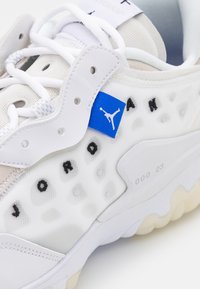 Weiße Sportschuh mit Mesh- und synthetischen Materialien, einem blauen Etikett und schwarzer Schrift. Strukturiertes Design entlang der Seite und eine Gummisohle.