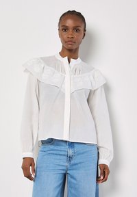 Weiße Bluse mit weitem Rüschensaum und gesticktem Detail auf der Brust, mit Knopfverschluss und langen Ärmeln mit gerafften Manschetten.