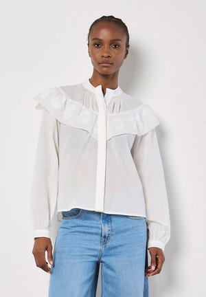 Weiße Bluse mit weitem Rüschensaum und gesticktem Detail auf der Brust, mit Knopfverschluss und langen Ärmeln mit gerafften Manschetten.