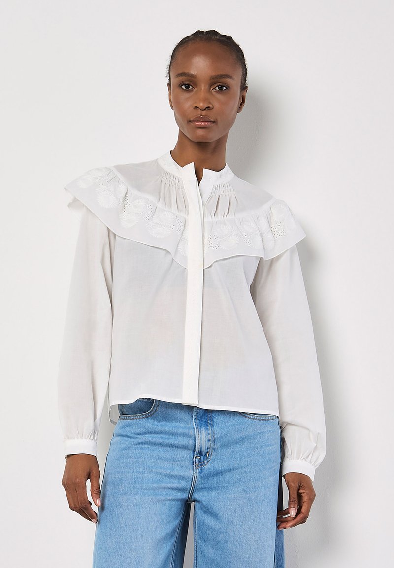 Weiße Bluse mit weitem Rüschensaum und gesticktem Detail auf der Brust, mit Knopfverschluss und langen Ärmeln mit gerafften Manschetten.