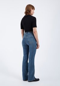 Kvinde med kort brunt hår står vendt mod højre iført en sort tætsiddende top og blå højttaljede jeans med sorte sko.