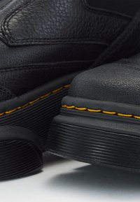 Gros plan sur des bottes noires texturées avec des semelles épaisses et une couture jaune le long du bord de la semelle, mettant en évidence les détails du matériau et de la construction.