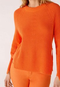 Femme portant un pull en maille côtelée orange vif à manches longues et un pantalon orange assorti, court à la taille.