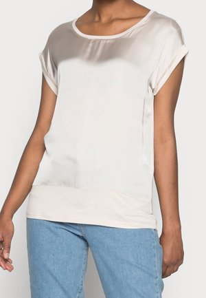 Vrouw draagt een losse, korte mouwen, licht beige satijnen blouse met opgerolde manchetten, gecombineerd met lichtblauwe spijkerbroeken.