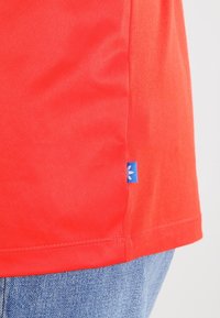 Röd bomull T-shirt med en slät yta, med en blå logotypapplikation vid sömmen och rak fåll. Avslappnad passform, korta ärmar.