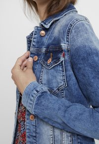 Denim jacket with a faded blue finish, copper-tone buttons, and two chest pockets. Features a pointed collar and visible stitching details.