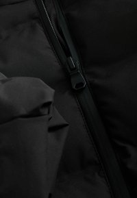 Tissu de veste rembourrée noire avec une fermeture éclair visible. Le matériau semble lisse et mat, avec un design épuré et minimaliste et des coutures dissimulées.