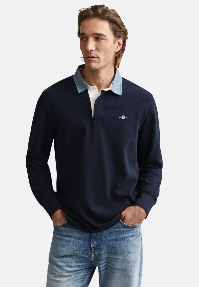 Marineblauw poloshirt met lange mouwen, een lichte denim kraag en een witte knoopsluiting, met een geborduurd logo op de borst. Draagt lichtblauwe spijkerbroek.