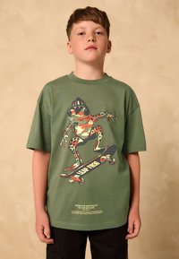 Zelena pamučna majica s grafikonom žabe na skateboardu s višebojnim naglascima. Kratki rukavi, opušteni kroj i ispisani tekst.