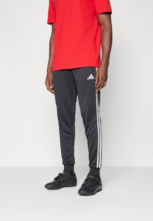 Svarta sportbyxor med vita sidostråk och en liten logotyp, i kombination med en röd bomulls t-shirt och svarta träningsskor.