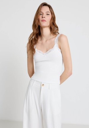 Jeune femme aux cheveux longs et ondulés portant un caraco blanc orné de dentelle et un pantalon blanc plissé taille haute, sur fond uni.