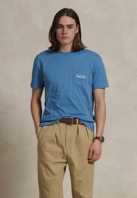 Svijetloplava pamučna majica s džepom na prsima i bijelim vezanim tekstom, u kombinaciji s bež nabranim hlačama i smešnim pojasom.